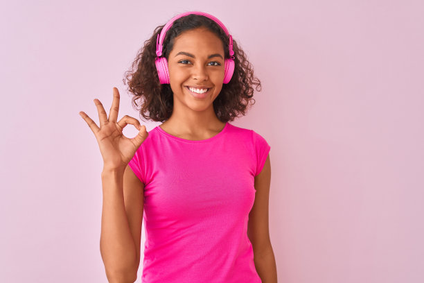 一名年轻的巴西女子在孤立的粉色背景下使用耳机听音乐，用手指做ok手势，非常棒的符号图片下载