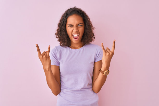 年轻的巴西女人穿着t恤站在孤立的粉红色背景大喊与疯狂的表情做摇滚符号与手举起。音乐明星。沉重的音乐概念。图片下载