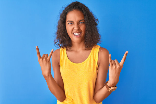 年轻的巴西女人穿着黄色的t恤站在孤立的蓝色背景大喊与疯狂的表情做摇滚符号与手举起。音乐明星。沉重的概念。图片下载