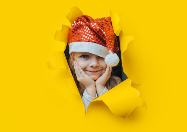等待圣诞礼物。一个漂亮的小女孩戴着一顶红色的圣诞老人帽，双手抱着头，从一个用黄色纸做的洞里往外看。圣诞节和新年的概念。副本的空间。撕裂的背景。图片下载
