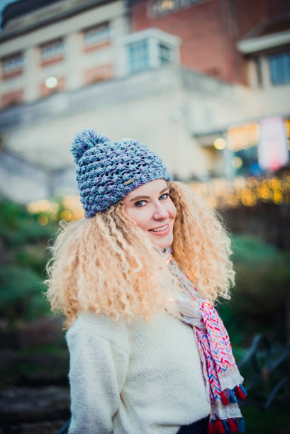 微笑的年轻卷曲的金发女人的肖像在温暖的白色自然毛衣和蓝色针织无檐帽与绒球看着相机与绿色公园的背景和散景抽象灯光。图片下载