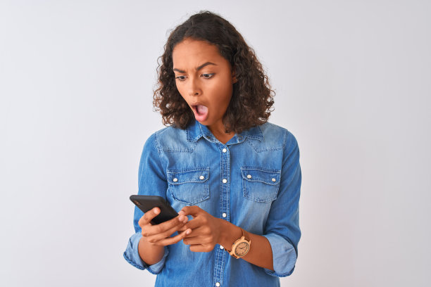 一名年轻的巴西女子使用智能手机站在孤立的白色背景下，惊恐地看着一张惊讶的脸，害怕和兴奋的恐惧表情图片下载