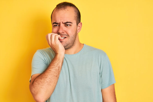 年轻男子穿着绿色休闲t恤站在孤立的黄色背景看起来紧张和紧张的手在嘴上咬指甲。焦虑的问题。图片下载