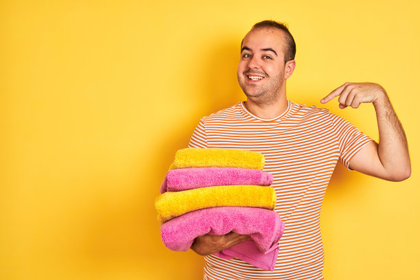 年轻的店主男子拿着折叠的毛巾站在孤立的黄色背景与惊讶的脸指向自己的手指图片下载