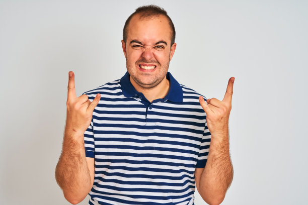 年轻人穿着休闲条纹马球站在孤立的白色背景大喊与疯狂的表情做摇滚符号与手举起。音乐明星。沉重的音乐概念。图片下载