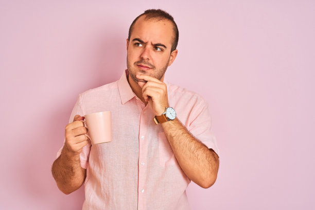 年轻人喝着咖啡，站在孤立的粉红色背景严肃的脸上思考着问题，非常困惑的想法图片下载