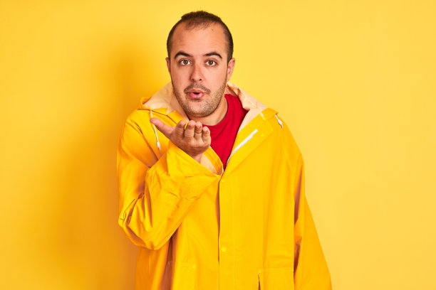 年轻男子穿着雨衣站在孤立的黄色背景，看着镜头飞吻与手在空中是可爱和性感的。爱的表达。图片下载