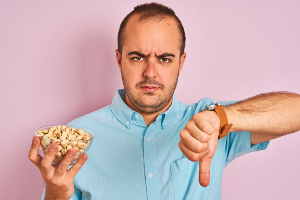 年轻男子拿着装有开心果的碗站在孤立的粉红色背景上，面带愤怒，这是表示不喜欢的负面信号，用大拇指朝下，拒绝的概念图片下载