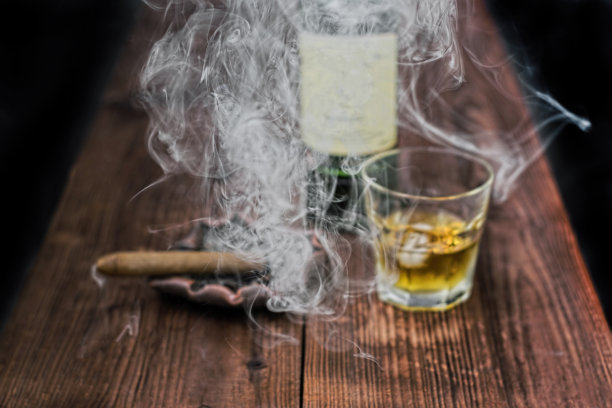 一张前景聚焦的雪茄烟雾的详细照片。背景是桌子，烟灰缸上放着一根雪茄，一杯加冰的威士忌和一个瓶子图片下载