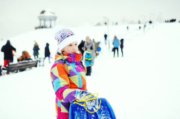 在雪坡的背景上，穿着彩色外套的小女孩手里拿着一个雪橇儿童背带。图片下载