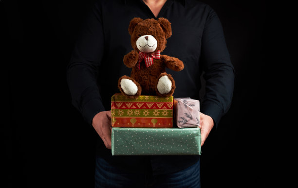 一名穿着黑色衬衫的成年男子拿着一个用红丝带和棕色泰迪熊绑着的蓝色方盒子图片下载