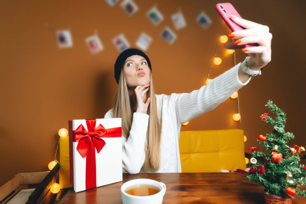 戴着美国帽子的漂亮女人在用智能手机自拍。圣诞树放在木桌上图片下载