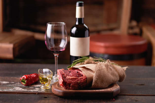 一瓶红酒和一杯，旁边是一块用羊皮纸和粗麻布裹着的陈年里贝，放在切菜板上，撒上豌豆，旁边是一碗植物油和红辣椒。烹饪牛排。景深浅图片下载