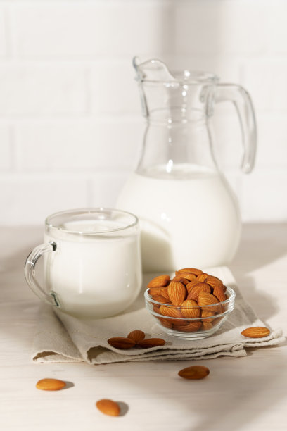 白色木桌上放着一个装有杏仁牛奶的玻璃瓶和大杯子，晨光透过窗户照进来。厨房里有一碗杏仁。图片下载