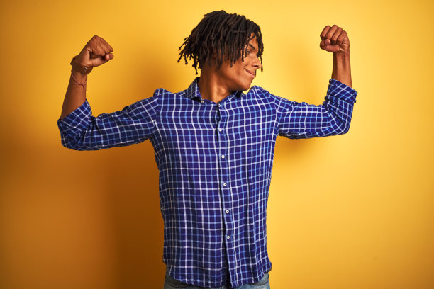 一个留着长发绺、穿着休闲衬衫的黑人站在孤立的黄色背景下，露出手臂上的肌肉，自豪地微笑着。健康的概念。图片下载