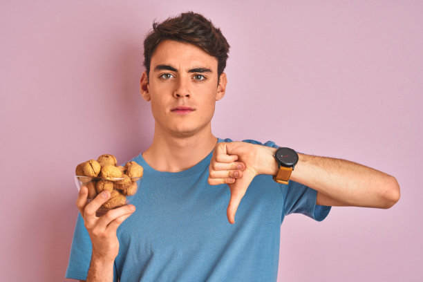 十几岁的男孩拿着一束天然核桃在粉红色孤立的背景与愤怒的脸，消极的迹象显示不喜欢拇指向下，拒绝概念图片下载