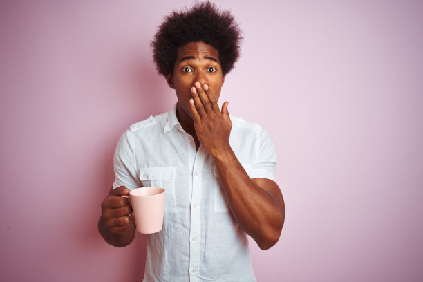 年轻的非洲裔美国人喝一杯咖啡站在孤立的粉红色背景覆盖嘴与手震惊为错误，表达恐惧，在沉默中害怕，秘密的概念图片下载