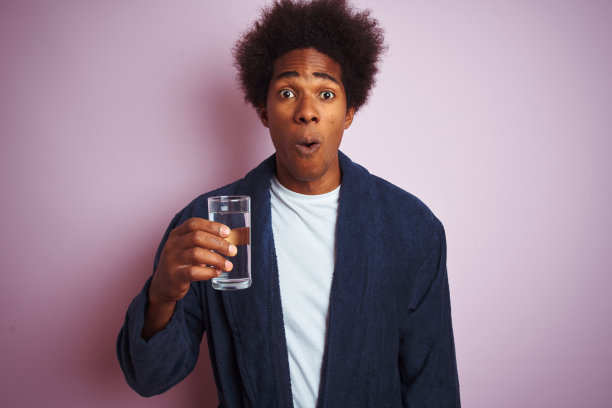 非裔美国人穿着睡衣，喝了一杯水在孤立的粉红色背景吓了一跳，吃惊的脸，害怕和兴奋与恐惧的表情图片下载