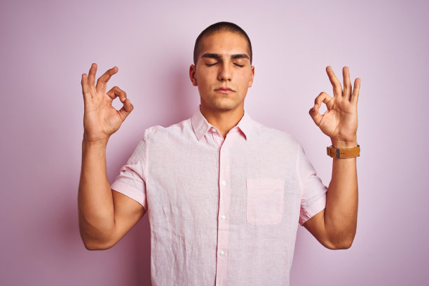 年轻英俊的男子穿着优雅的衬衫在粉红色孤立的背景放松和微笑，闭上眼睛做冥想手势与手指。瑜伽的概念。图片下载