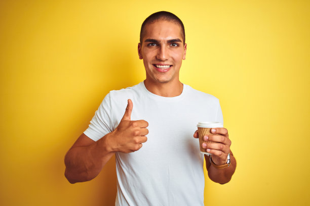 年轻英俊的男子喝了一杯带走的咖啡在黄色孤立的背景愉快地与大微笑做ok的手势，拇指与手指竖起，优秀的手势图片下载