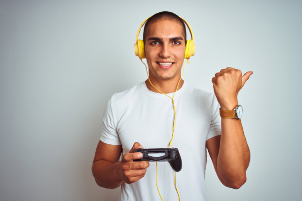 年轻英俊的男子玩电子游戏使用耳机白色孤立的背景指向和显示与拇指向上的一边与快乐的脸微笑图片下载