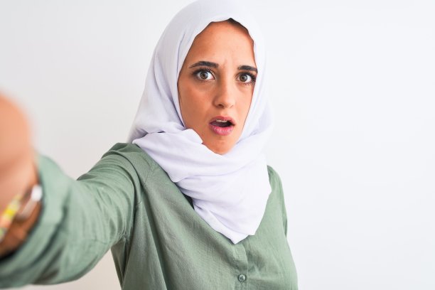 年轻的阿拉伯妇女戴着头巾自拍照片在孤立的背景下害怕在震惊与惊讶的脸，害怕和兴奋与恐惧的表情图片下载