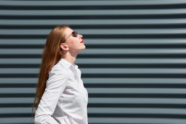 红发年轻迷人的女商人戴着太阳镜和白衬衫，涂着红色的嘴唇看着太阳，享受这一刻。灰色金属面板背景。图片下载