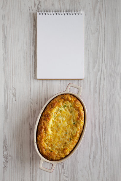 自制切达玉米布丁砂锅，白色木质表面上的空白记事本，俯视图。平躺，俯视图，从上面。副本的空间。图片下载