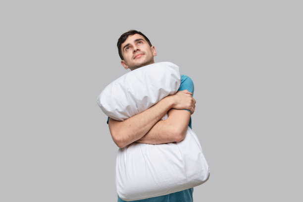 年轻的深色头发的男人抱着白色的枕头在灰色的背景上孤立地做梦。睡觉的概念。图片下载