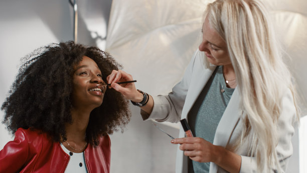 后台的照片拍摄:化妆师应用口红化妆美丽的黑色模特与郁郁葱葱的卷发。时尚杂志工作室拍摄准备。专业设备工作室图片下载