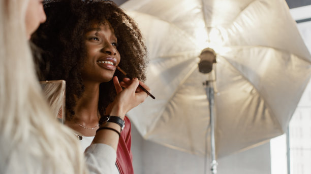 照片拍摄后台:化妆师为美丽的黑人女孩涂抹口红化妆。专业模特正在为时尚杂志拍摄摄影。图片下载