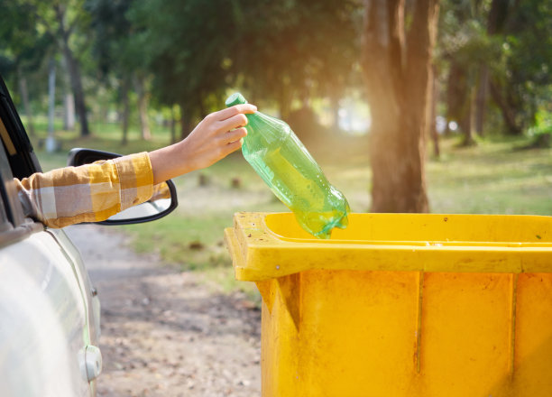 女手把一个绿瓶子扔到公园路边的黄色垃圾桶里。图片下载