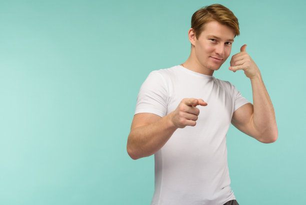 打电话给我。英俊的年轻微笑的男子看着相机和手势手机近脸，而站在蓝色的背景图片下载