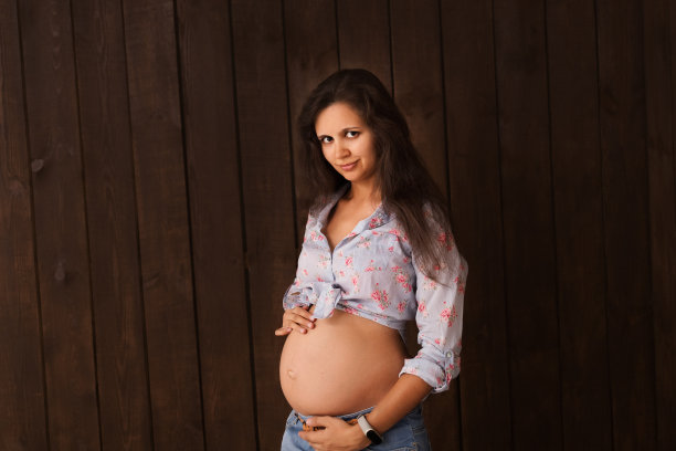 穿着牛仔裤短裤和蓝色衬衫的孕妇双手放在深棕色的背景上。怀孕、分娩、准备和期望概念。美丽柔情的怀孕照片图片下载