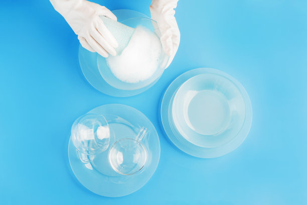 女性双手在珍珠白色手套洗涤美丽的浅蓝色玻璃盘子和杯子在时尚的蓝色背景。洗碗的概念。背景为您的设计。平的风格。图片下载