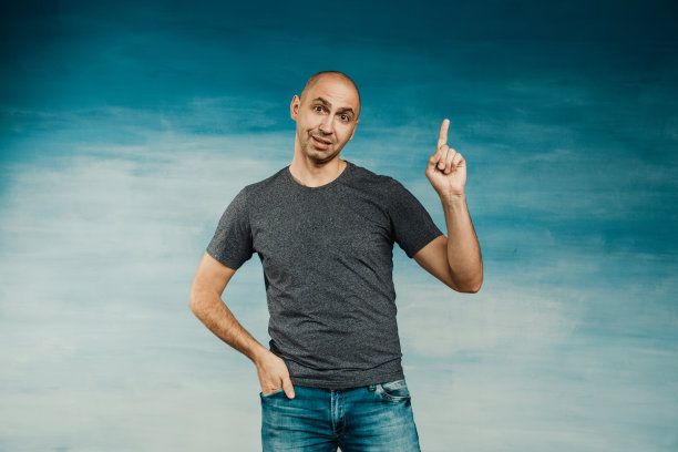 蓝色背景下，一个身穿灰色t恤的年轻秃顶男子惊讶地看着摄像机，举起了他的索引。图片下载