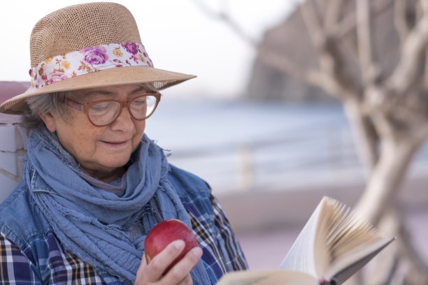 一位老年妇女享受退休生活的方式是坐在海滩边看书，吃苹果。水面上的地平线。海洋景观。一个只有戴眼镜和帽子的人图片下载