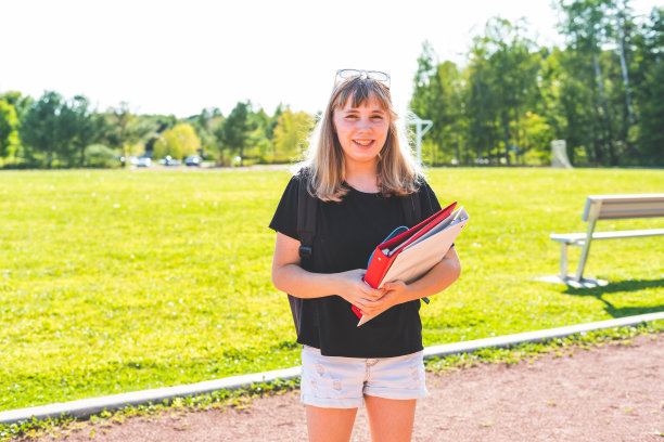 快乐微笑的少女学生站在高中和足球场前，拿着活页夹，戴着太阳镜图片下载
