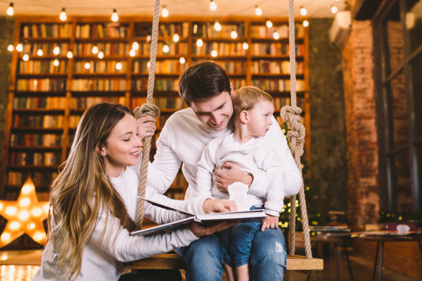 在平安夜的舒适黑暗的客厅里，有婴儿的家庭在家里的秋千座位上放松。家人玩跷跷板。和家人和孩子在冬天的夜晚。客厅里的家庭读物图片下载