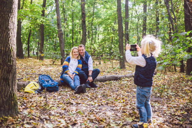旅游、旅游、远足、科技、家庭理念。快乐的孩子让照片的父母在森林里。爸爸妈妈摆姿势拍照，女儿用手机拍照。森林里的年轻游客家庭图片下载