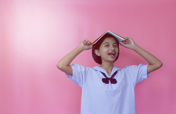 快乐美丽的年轻亚洲女孩拿着书与制服，粉红色的背景。图片下载