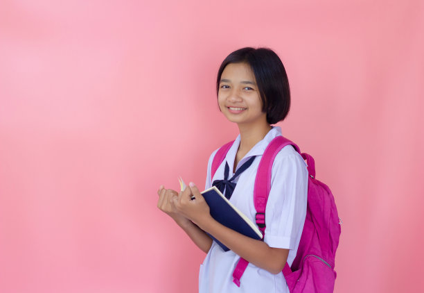 快乐美丽的年轻亚洲女孩拿着书与制服，粉红色的背景。图片下载