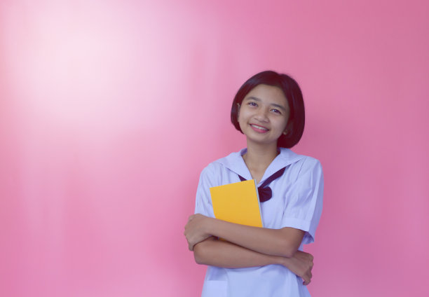 快乐美丽的年轻亚洲女孩拿着书与制服，粉红色的背景。图片下载