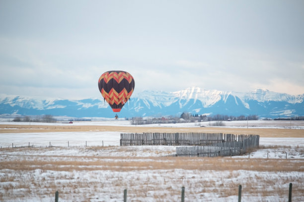 在一个阴天的冬天，一个热气球在落基山脉旁边的空旷的田野上飞行。图片下载
