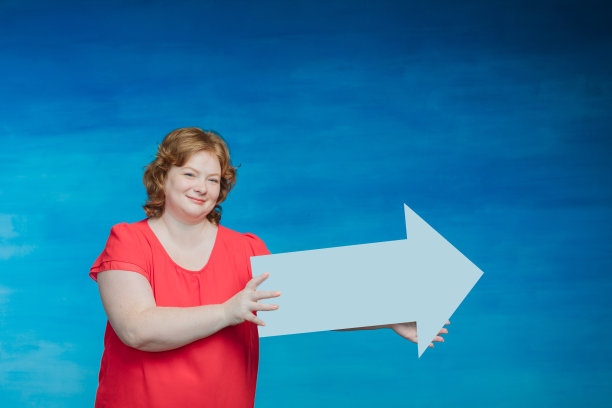 年轻迷人的丰满女人举着一个箭头指向一边的蓝色背景。图片下载