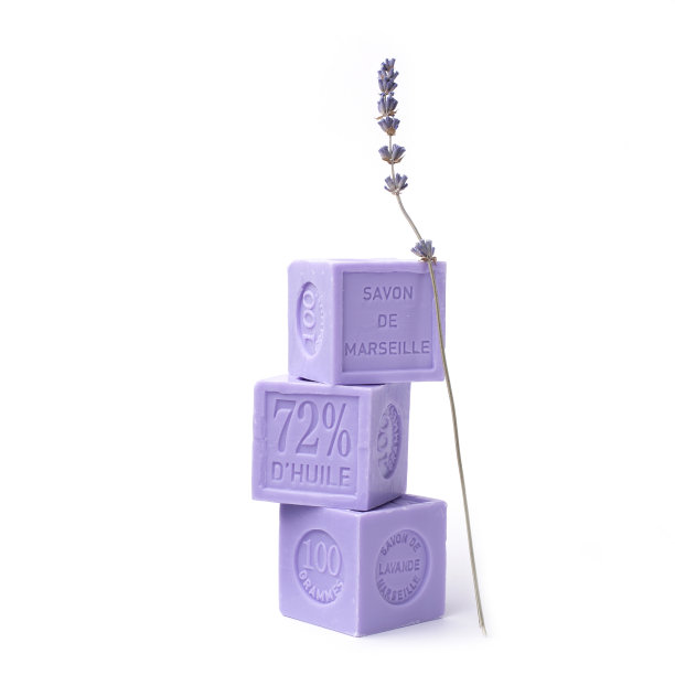 三种淡紫色的天然薰衣草皂，每一种都有法语铭文:马赛薰衣草皂，100克，一层一层排列，白色桌子上放着薰衣草花。图片下载