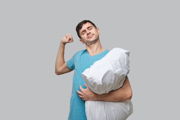 年轻的黑发男子伸展着白色的枕头孤立在灰色的背景。图片下载