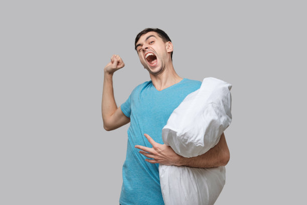 年轻的黑发男子伸展着白色的枕头孤立在灰色的背景。图片下载