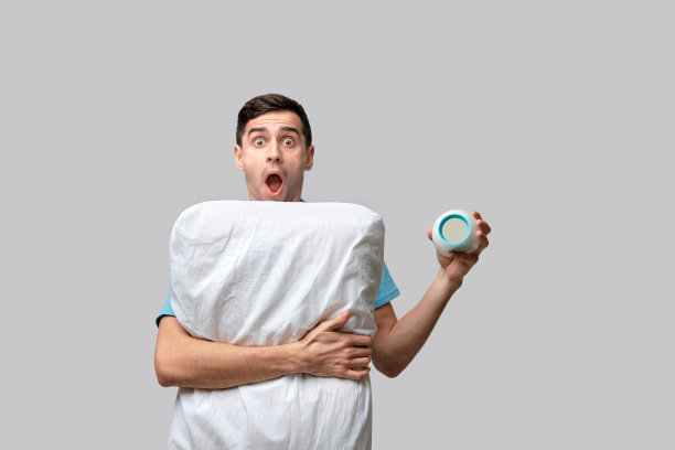 年轻的深色头发的男人是沮丧的抱着闹钟和白色的枕头孤立在灰色的背景图片下载