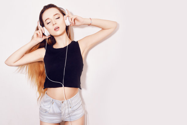 时尚靓丽的酷女孩戴着耳机，在蓝色背景上听着音乐。美丽的年轻少女，长发，跳舞图片下载